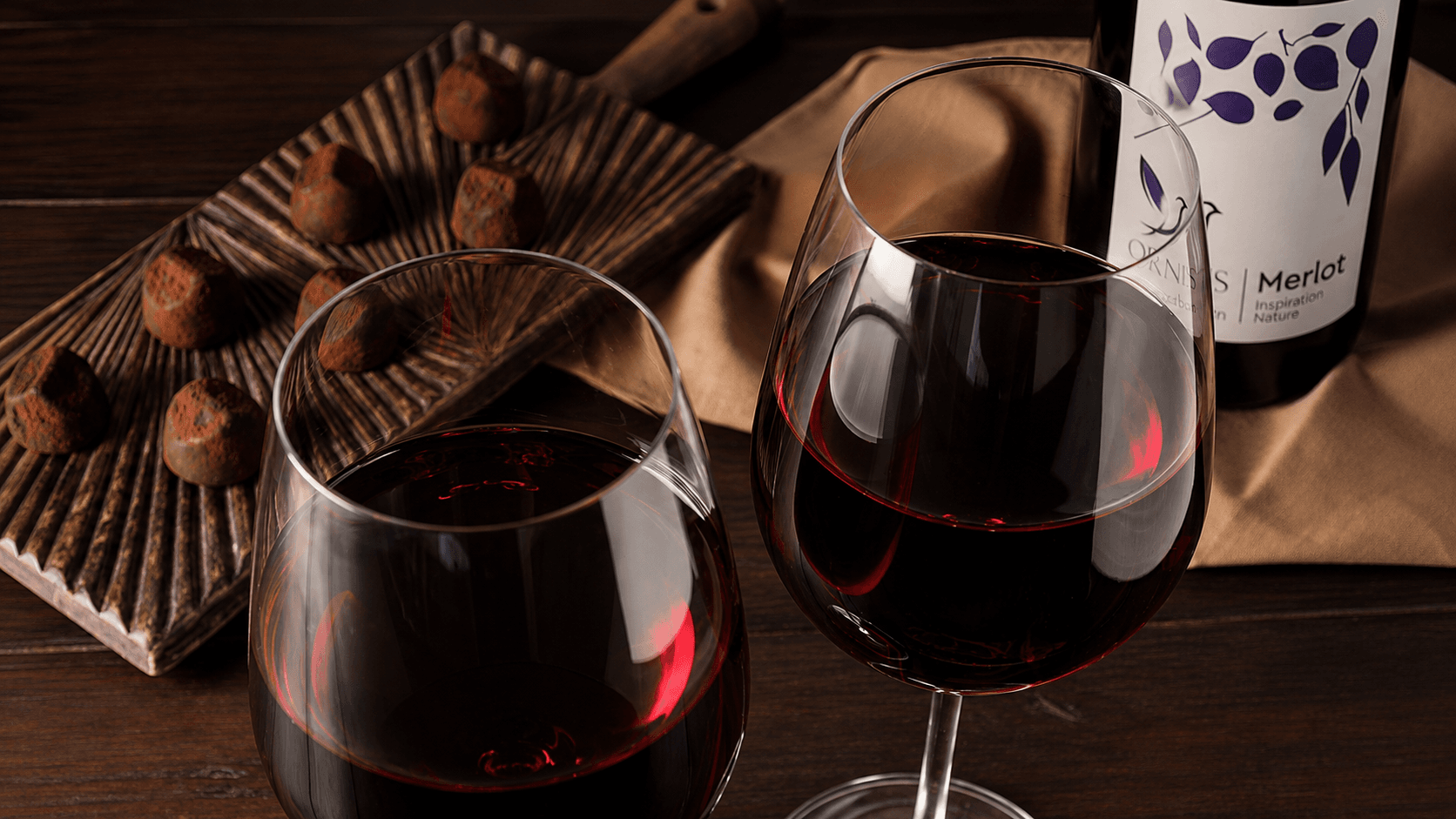 Бутылка ORNIS Merlot Inspiration Nature, два бокала красного вина и шоколадные конфеты на деревянном столе.